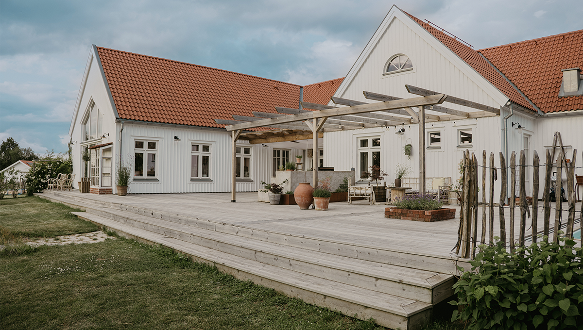 Stor träaltan med pergola utanför ett vitt hus med röda takpannor. Altanen sträcker sig längs husets fasad och är möblerad med sittplatser och planteringar.